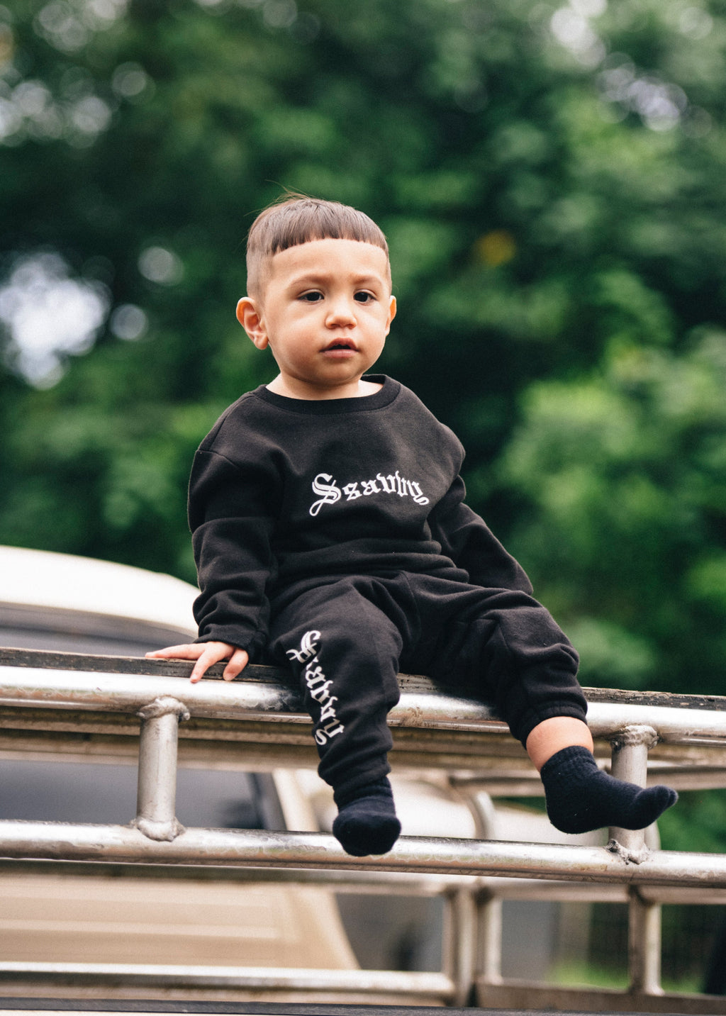 Child wearing a black outfit with visible branding, sitting on a metal structure with greenery in the background
